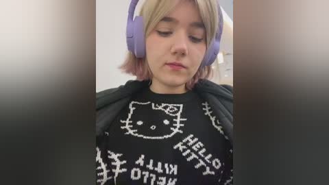 Media: Video of a young woman with short blonde hair, wearing large purple headphones, a black \"Hello Kitty\" shirt, and a black hoodie, looking introspective indoors.