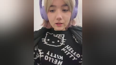 Media: A video of a young woman with fair skin and blonde hair wearing large purple headphones, a black \"Hello Kitty\" sweatshirt, and a black scarf, indoors with blurred background.