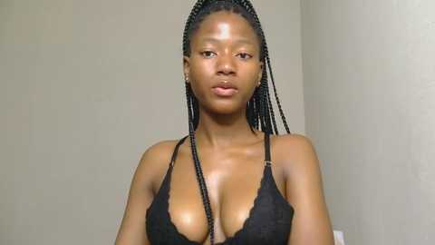 Media: Video of a young, slender Black woman with medium-brown skin, wearing a black lace bra, showcasing her medium-sized breasts. She has neat, braided hair and a neutral expression, set against a plain, beige wall background.