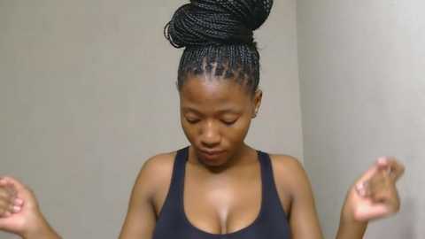 Media: Video of a dark-skinned African woman with medium-sized breasts, wearing a black sports bra, her hair styled in tight braids with a large bun. She stands indoors, hands raised in a yoga pose, against a plain beige wall.