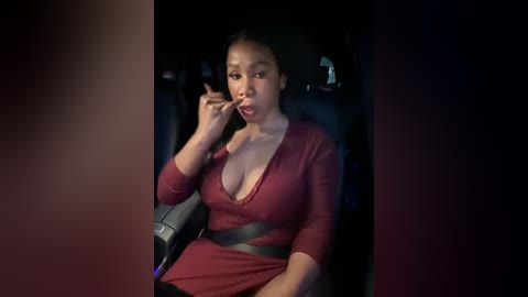 Media: Video of a young Black woman with medium skin tone, dark hair tied back, wearing a low-cut red dress, seated in a dimly lit car, holding a cigarette to her lips, dim background.