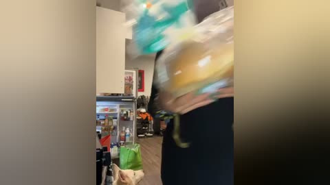 Media: Video of a blurred figure in a colorful, motion-blurred shirt and black pants, holding a green bag, walking into a brightly lit, modern apartment with a white refrigerator and cluttered kitchen.