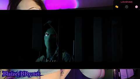 Media: Video of a woman in a dark, greenish hooded costume, partially illuminated by a purple light, standing in a dimly lit room. The image includes a watermark reading \"BlazeyBrynet.net\" at the bottom.