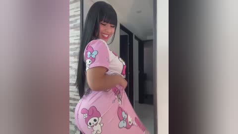 Media: Video of a Latina woman with long black hair and medium skin tone, wearing a pink My Melody dress, posing in a modern, minimalist hallway.