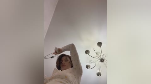 Media: Video of a woman with short brown hair, wearing a white sweater, reaching up to adjust a chandelier in a white room.