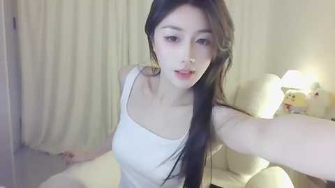 Media: A video of an Asian woman with long black hair, fair skin, and a slim figure, wearing a white tank top, taking a selfie in a softly lit, minimalist room with cream-colored curtains and a white lamp.