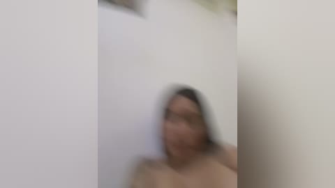 A blurred, out-of-focus video of a naked woman with shoulder-length brown hair, possibly of Asian descent, against a white wall. The image lacks clarity and detailed features.