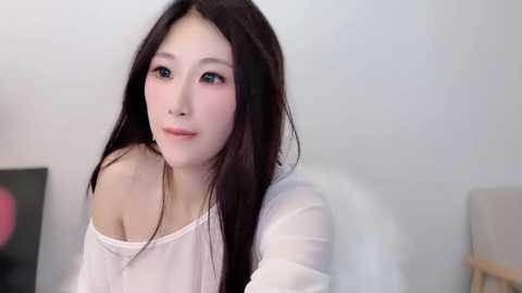 Media: Video of a young Asian woman with long, straight black hair, fair skin, and a petite frame. She wears a white off-shoulder top. Background features a white wall and blurred furniture.