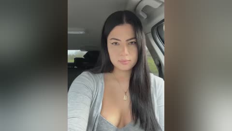 Media: Video of a young woman with long black hair, fair skin, and a medium build, wearing a grey cardigan over a low-cut grey top, seated in a car.