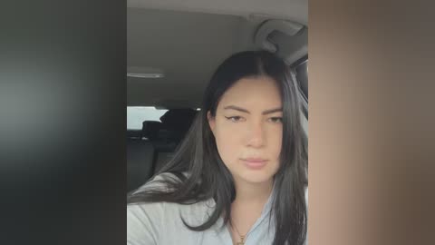 Media: A video of a young woman with long black hair, light skin, and a neutral expression, wearing a light blue shirt, sitting in a car with a blurred background of a dark interior.