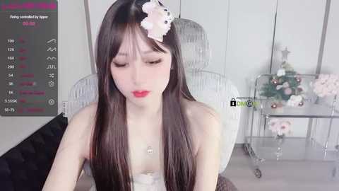 Media: Video of a young Asian woman with long, straight black hair, adorned with a pink flower accessory, sitting in a modern, minimalist room. She wears a strapless white dress and has a serene expression.