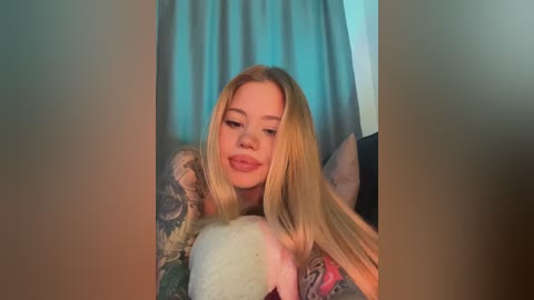 A video of a young, fair-skinned, blonde woman with a slight smile, wearing a floral-patterned shirt, resting her head on a large, fluffy white pillow. The background features teal curtains and a muted room.