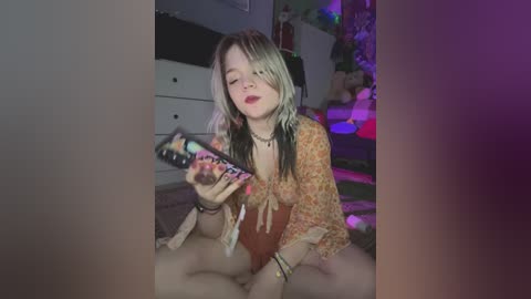 Media: A young woman with long, blonde hair, wearing a floral top and a necklace, sits on a bed, holding a phone, with a blurred background of a room lit by purple and green lights.