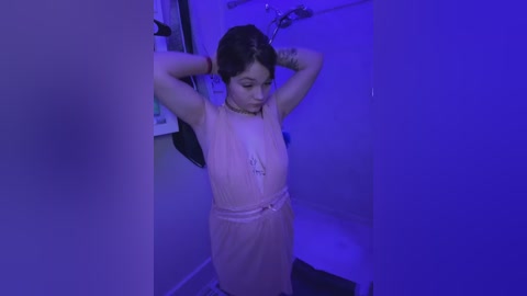 Media: Video of an Asian woman with fair skin, wearing a sleeveless peach dress, standing in a dimly lit bathroom with blue lighting. She has dark hair tied back, arms raised, and is looking downward.