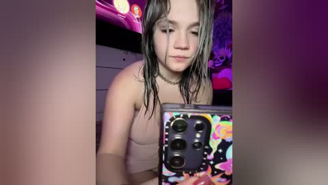 Media: Video of a young woman with wet, dark hair, wearing a beige tank top, taking a selfie in a dimly lit room with colorful lights and decorations in the background.