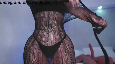 Media: A video of a woman in black fishnet lingerie, standing in a dimly lit room. Her physique is slender, with visible breasts and a high-cut thong. A blurred figure lies on a bed in the background.