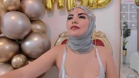 Media: Video of a fair-skinned woman in a sheer white dress with a silver hijab and a gold crown, surrounded by metallic balloons, seated on a red and gold throne.