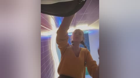 Media: Video of a blonde woman in a mustard-yellow sweater, mid-air, reaching for a black shoe in a tiled hallway with blue and purple lighting.