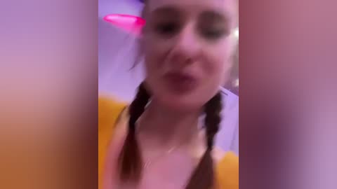 Media: Video of a young woman with pale skin, dark hair styled in braids, wearing a yellow top, and a pink, heart-shaped nose filter. Background is blurred and purple-hued, creating a soft, dreamy atmosphere.