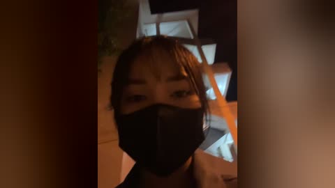 Media: Video of an Asian woman with light skin, wearing a black face mask, brown hair, and a white top, standing in a dimly lit room with a staircase and green plants in the background.