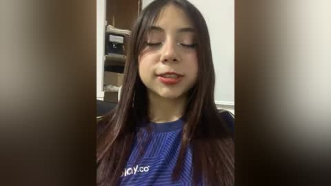 Media: A video of a young Latina woman with long, straight, dark hair, wearing a blue shirt, standing in a salon with a wooden cabinet and shelves in the background.