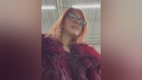 Media: Video of a woman with short, bright orange hair, wearing black-rimmed glasses and a dark fur coat, standing in an industrial setting with a tiled ceiling and fluorescent lights.