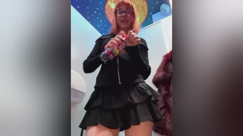 Media: A video of a pale-skinned woman with red hair and glasses, wearing a black jacket and short skirt, holding a spray bottle in a brightly lit room with a starry night ceiling mural.