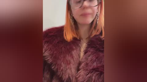 Media: Video of a woman with shoulder-length, straight, orange hair, wearing glasses and a plush, maroon fur coat with a high collar. Background is a blurred, reddish-brown gradient.