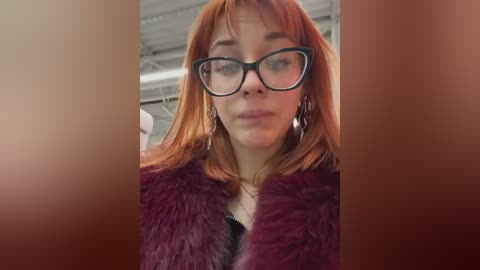 Media: Video of a fair-skinned woman with red hair, wearing black-rimmed glasses and a maroon faux-fur coat, standing in a warehouse setting.