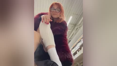 Media: Video of a woman with shoulder-length orange hair, wearing glasses, a burgundy fur coat, white sweater, and black boots, sitting on a chair in a well-lit room with white ceiling and shelves in the background.