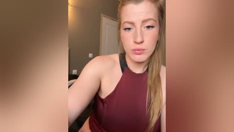 Media: Video of a fair-skinned, blonde woman with light makeup, wearing a sleeveless maroon top, making a kissy face, in a beige-walled room.