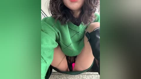 Media: A video of a woman with shoulder-length curly hair, wearing a green knitted sweater, black thigh-high stockings, and a pink vibrator, seated on a grey carpeted floor.