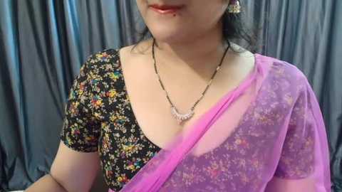 Media: Video of a woman with medium skin tone, wearing a black floral blouse and a sheer pink sari with a floral pattern. She has a delicate gold necklace and pearl earrings. The background features dark grey drapes.