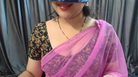 Media: Video of a South Asian woman in a pink saree with floral patterns, black floral blouse, and a blue face mask, smiling, against gray drapes.