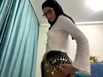 Media: A video of a slim woman with a medium skin tone and black hijab, wearing glasses and a tight, white textured top. She is posing with her hand on her hip, wearing a black skirt with gold sequin accents.