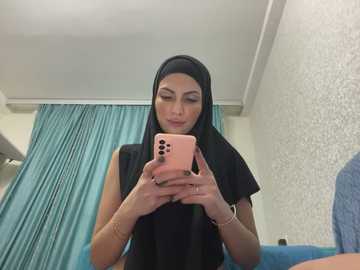 Media: Video of a light-skinned woman with long black hair and a black hijab, taking a selfie in a hospital room. She wears a sleeveless black top, with teal curtains and white textured wallpaper in the background.