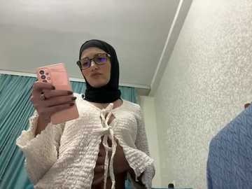 Media: Video of a young woman with a medium complexion, wearing a black hijab, glasses, and a cream-colored tie-front blouse, taking a selfie in a bedroom with teal curtains and white textured wallpaper.