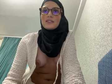 Media: Video of a light-skinned woman with glasses, black hijab, and partially unzipped cream cardigan revealing her bare breasts. She stands in a room with a teal curtain and textured white wall.