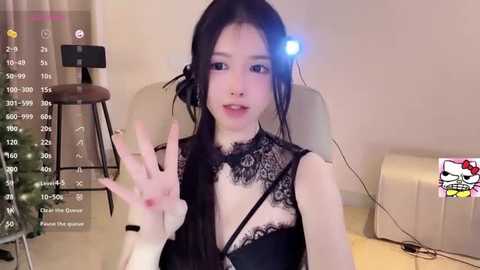 Media: Video of an East Asian woman with long black hair, wearing a black lace top, sitting in a chair. She waves with her right hand, in a livestream setting, with a webcam and a \"Hello Kitty\" sticker on a white wall.