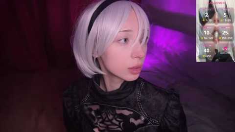 Video of a young woman with short, platinum blonde hair, wearing a black, lace-up, gothic-inspired outfit, set against a dimly lit, purple-hued background.