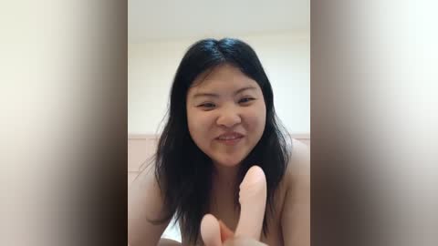 Media: Video of an East Asian woman with long black hair, fair skin, and a small nose, making a peace sign with her fingers, wearing a light-colored top, indoors with a blurred background.