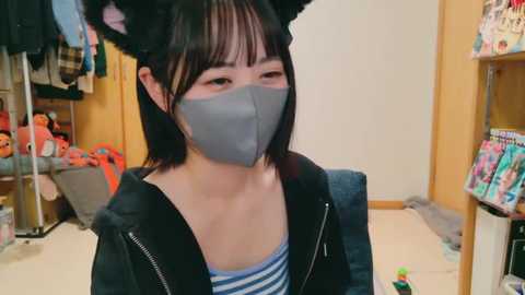 Media: Video of an Asian woman with short black hair, wearing a grey face mask, black hoodie, and striped shirt, in a cluttered room with stuffed animals and toys, and a wooden shelf in the background.