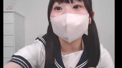 Media: Video of an Asian woman with long black hair and a white sailor uniform, wearing a white surgical mask, indoors with a white dresser in the background.