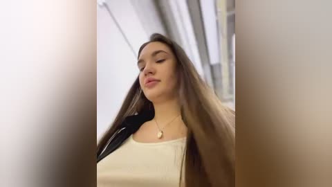 Media: Video of a young woman with long brown hair, wearing a white top and black jacket, standing in an indoor corridor with blurred background.