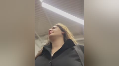 Media: Video of a woman with fair skin, long blonde hair, and a black blouse, looking up. She is in a modern, industrial-looking room with a high ceiling and fluorescent lights.