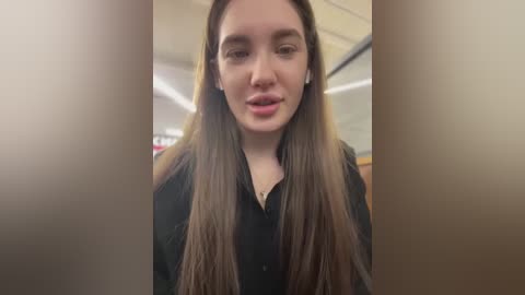 Media: A video of a young woman with long brown hair, fair skin, and a neutral expression, wearing a black blouse, taken indoors with a blurred background.