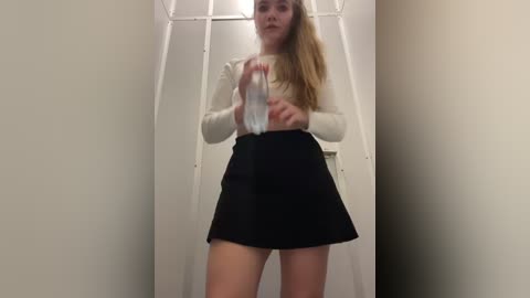 Media: Video of a young blonde woman in a white long-sleeve top and black skirt, holding a plastic bottle, inside a white cubicle with a metal frame.