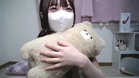 Media: A video shows an Asian woman with long dark hair, wearing a white mask and a white shirt, hugging a plush bear. The background features a pink curtain, a white shelf with stuffed animals, and a checkered bag.