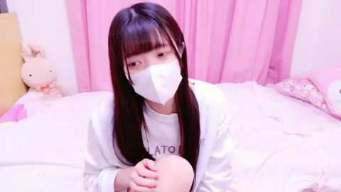 Media: A young Asian woman with long, straight black hair and bangs, wearing a white mask, white sweater, and white gloves, kneels on a bed with pink linens and pastel decor.