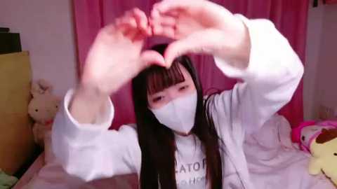 Media: Video of a young Asian woman with long black hair, wearing a white face mask, creating a heart shape with her hands in a bedroom with pink curtains and stuffed animals.